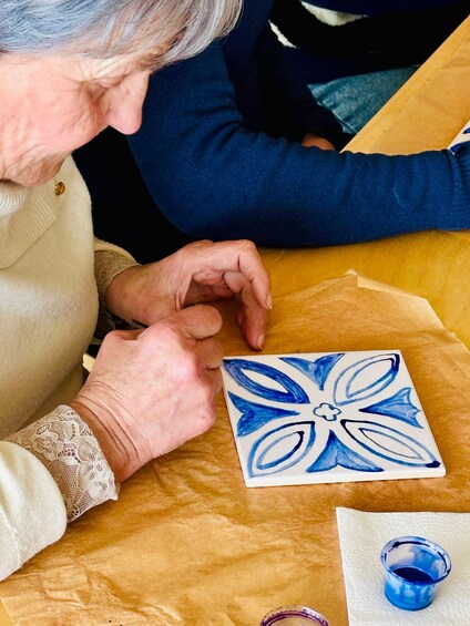 An elderly person painting a blue and white floral design on a ceramic tile. 4