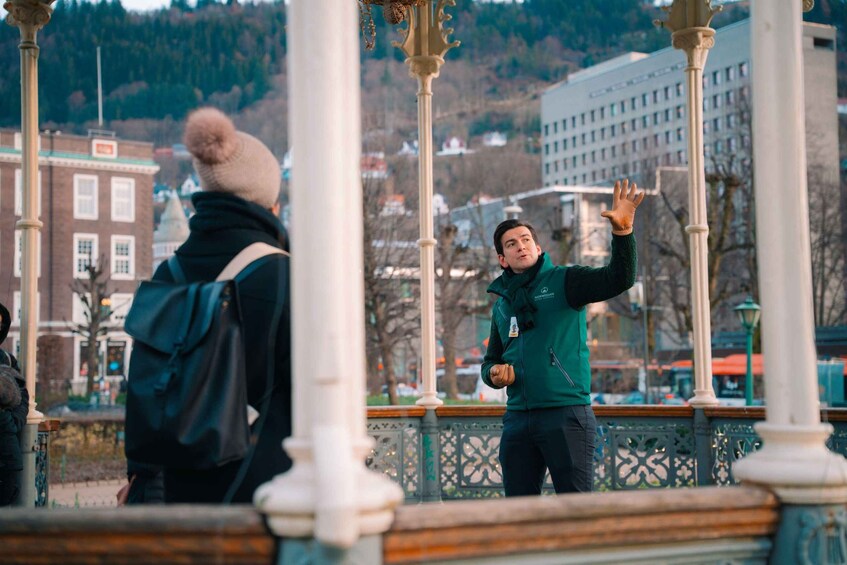A person in a green jacket standing on a bridge with ornate railings, a cityscape in the background, and another person in a black jacket with a fur-lined hood. 5
