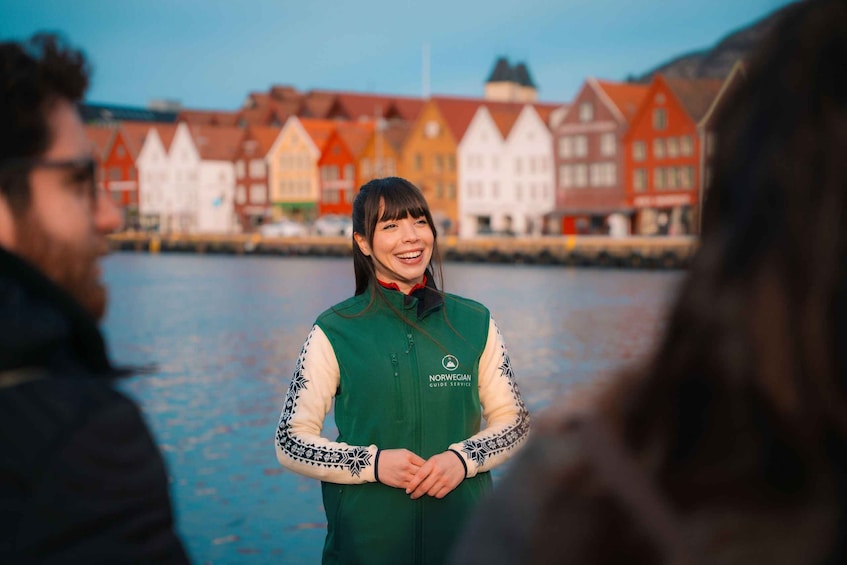 A person in a green vest with Nordic Guide Service logo, standing by a waterfront with colorful buildings in the background. 1