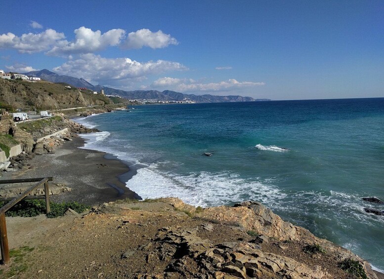 A coastal landscape with rocky shores, a clear blue sea, and distant mountains under a partly cloudy sky. 2