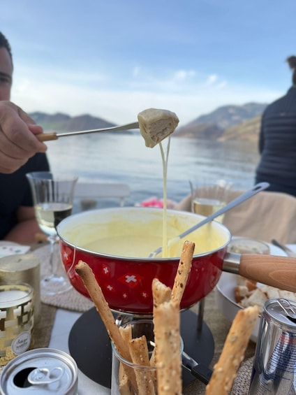 A person dipping a breadstick into a pot of melted cheese, with a scenic lake view in the background. 2