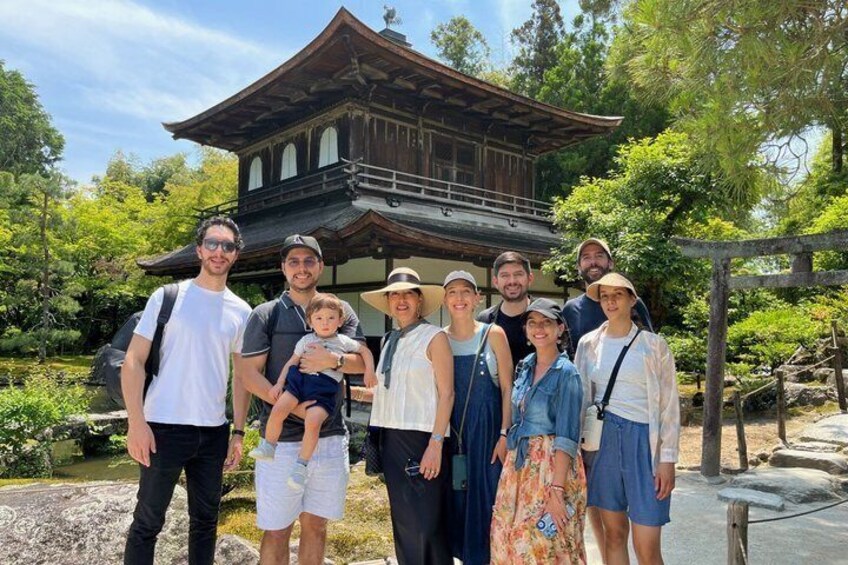 A group of people in front of a traditional wooden building with a tiled roof, surrounded by greenery. 1