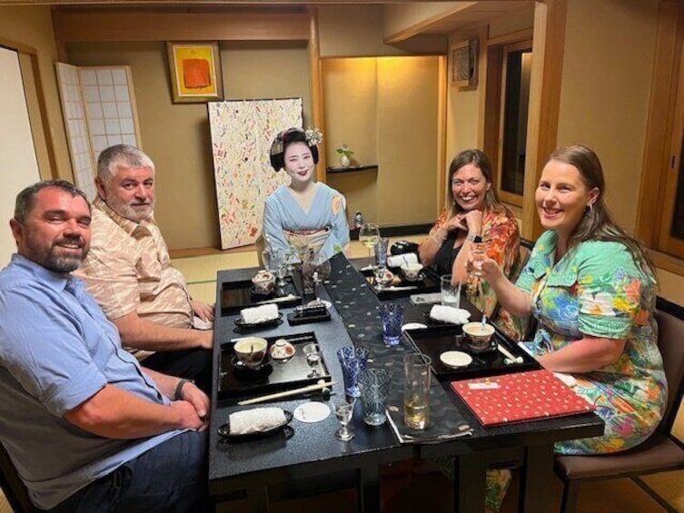 A group of people seated around a table with traditional Japanese dishes, a geisha, and a painting on the wall. 3