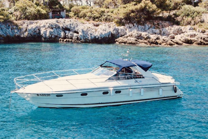 A white yacht with the registration number B60036 on calm blue waters, surrounded by rocky cliffs and sparse vegetation. 1