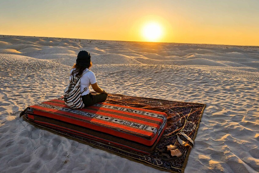 A person sitting on a patterned rug in a desert with sand dunes and the sun setting in the background. 3