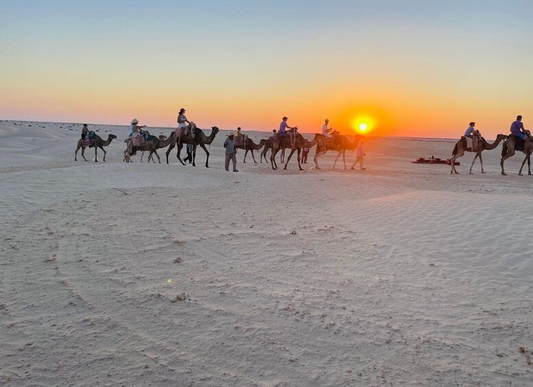 A group of people riding camels across a desert landscape during sunset. 1
