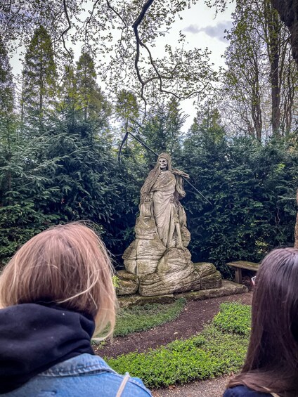 A statue of a skeletal figure with a scythe, surrounded by dense foliage and a stone bench. 4