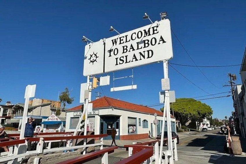 A welcome sign for Balboa Island, a small building, and a street with vehicles. 1