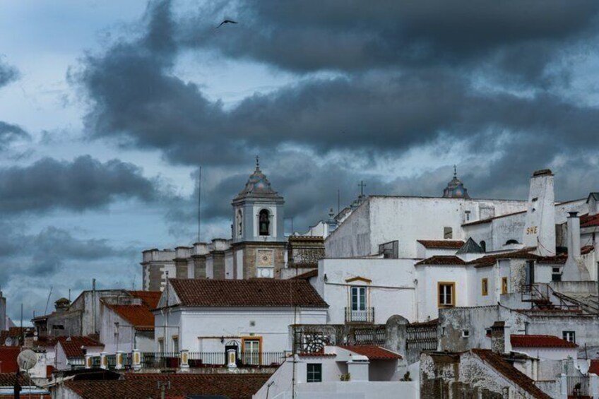A historic town with white buildings, a prominent church with a bell tower, and a cloudy sky. 3