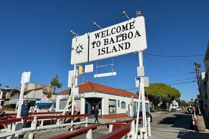 A welcome sign to Balboa Island, a small building, and a street with vehicles. 1