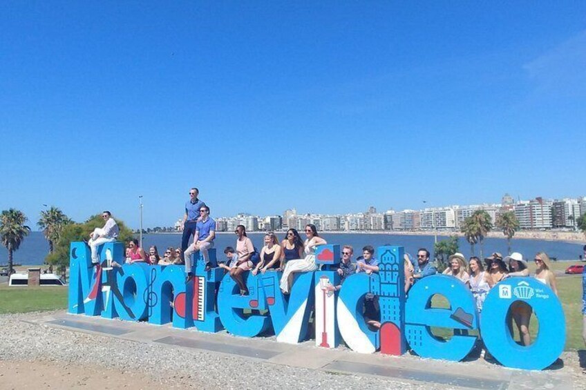 Eine Gruppe von Personen posiert vor einem großen, blauen Schild mit dem Wort 'Montevacio' und verschiedenen Symbolen. 1