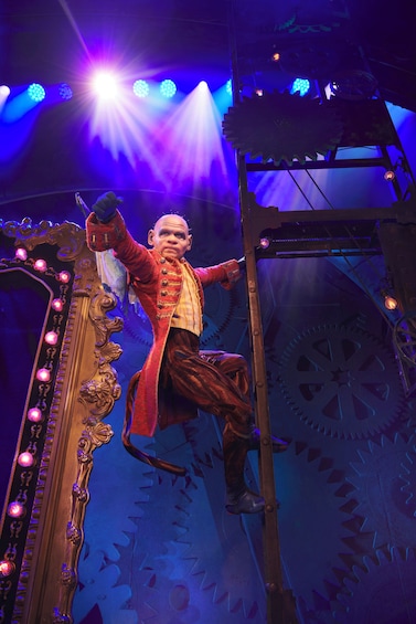 A person in a red coat climbing a ladder in a theater with a large mirror and mechanical gears. 4