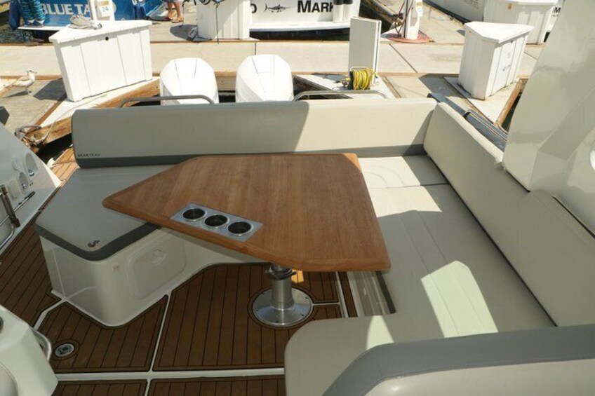 A boat's deck with a wooden table, white cushioned seating, and a white cooler. 3