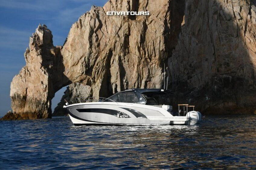 A white motorboat with a cabin, anchored near a rocky coastline with a large rock formation in the background. 1