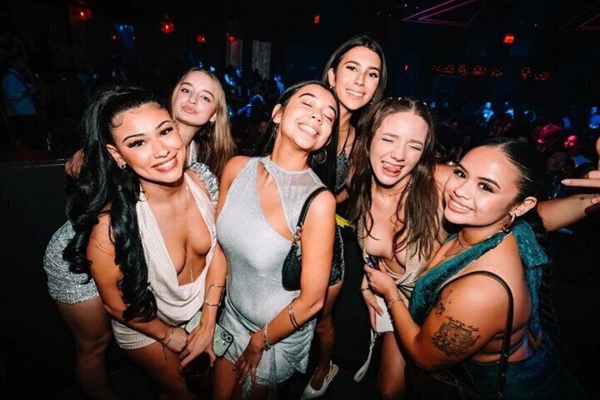 A group of people at a social event, possibly a party or club, with a dimly lit background and a crowd in the distance. 2