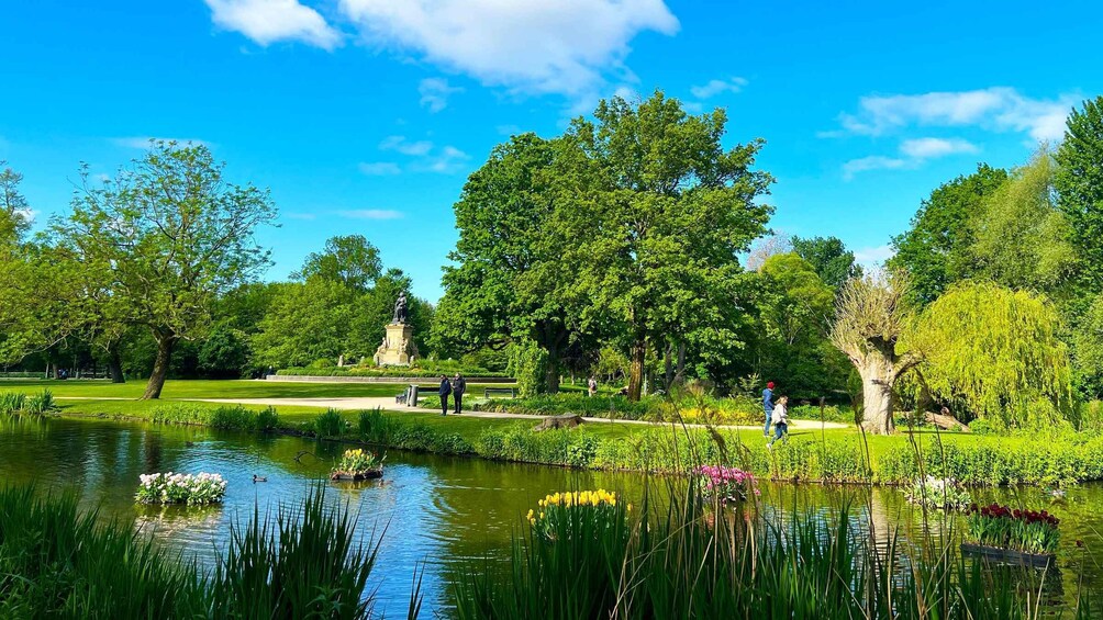 A park with a pond, trees, a statue, and people walking. 1
