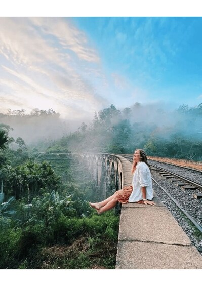 A person sitting on a concrete ledge by a railway track, with a bridge and lush greenery in the background. 3