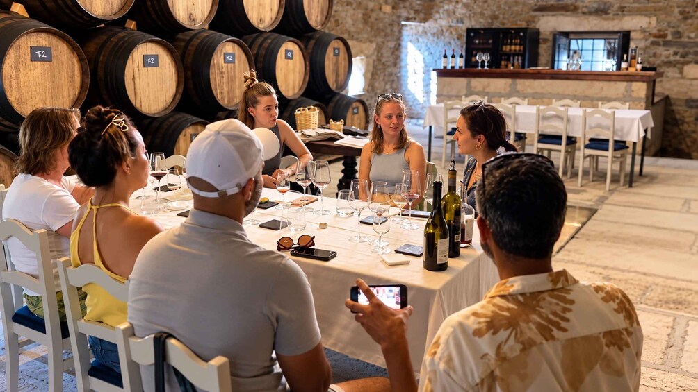 A group of people seated at a table with wine glasses and bottles, in a setting with wooden barrels in the background. 2