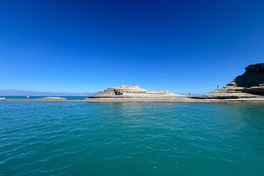 A coastal landscape with clear blue water, a rocky outcrop, and a distant lighthouse. 3