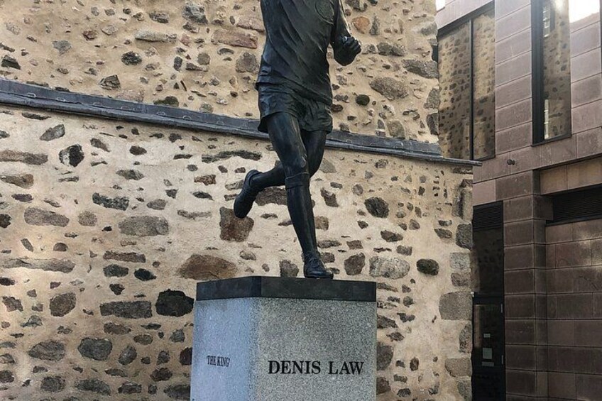 A statue of a man in motion, labeled 'DENIS LAW,' on a pedestal against a stone wall. 5