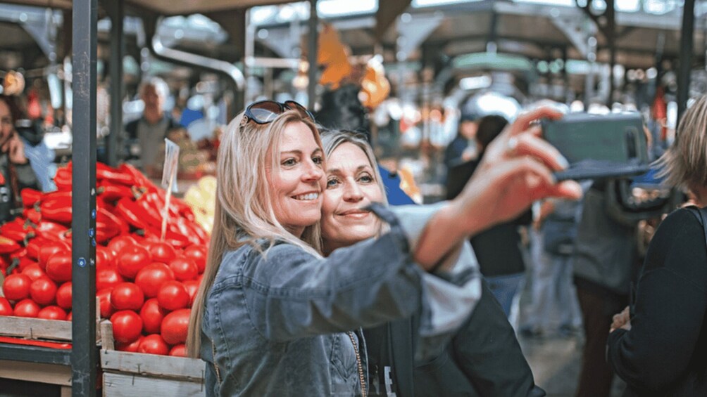 Two people taking a selfie at a market with tomatoes in the foreground. 1