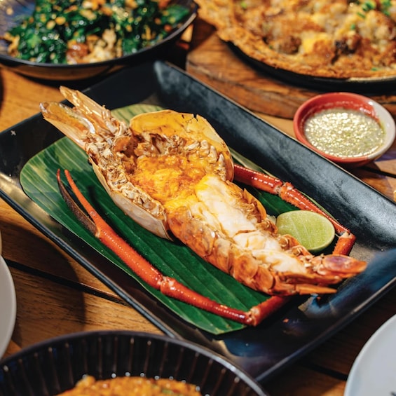 A large lobster on a banana leaf, with a lime wedge and red chili pepper on a black tray. 3
