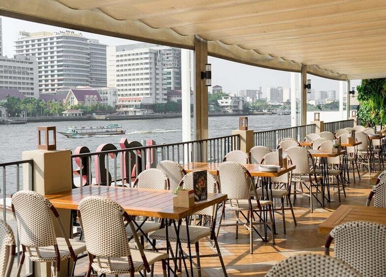 An outdoor restaurant with tables and chairs, overlooking a river with buildings and a boat in the background. 1