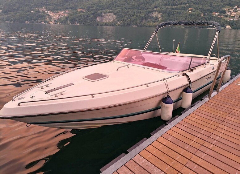A white motorboat docked at a wooden pier with a calm lake and a hillside in the background. 2