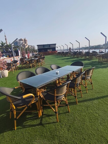 An outdoor dining area with a long table and chairs, surrounded by artificial grass, with a view of the water and buildings in the background. 2