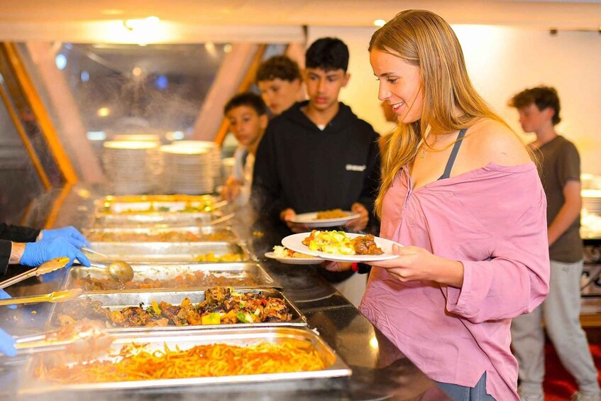 A buffet line with various dishes, a person serving food, and other individuals in the background. 3
