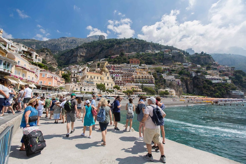 A coastal town with people walking along the promenade, overlooking a bay with boats and a mountainous backdrop. 1