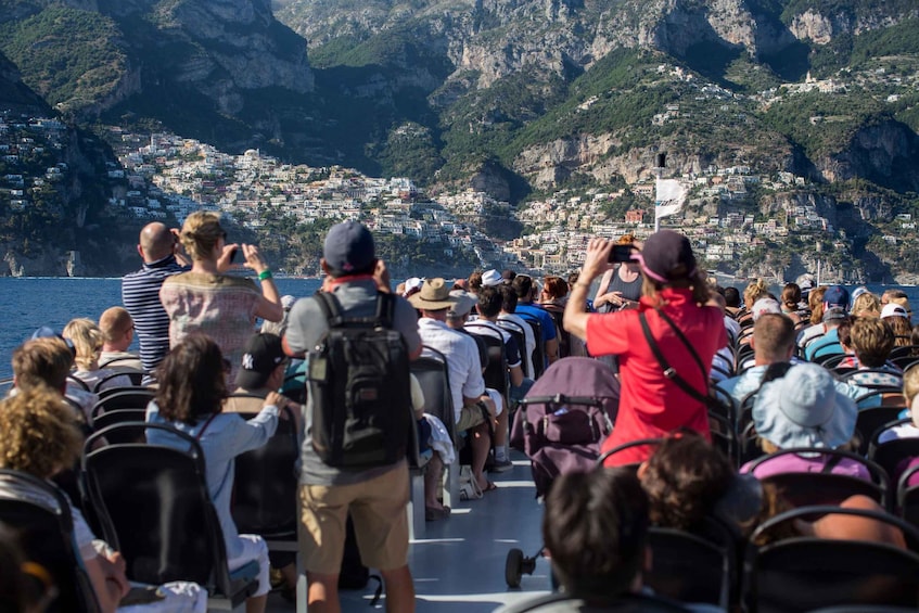A group of people on a boat, facing a coastal town with buildings on the cliffs. 3