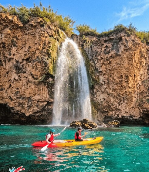 A waterfall in a rocky landscape with kayakers in a clear blue sea. 3