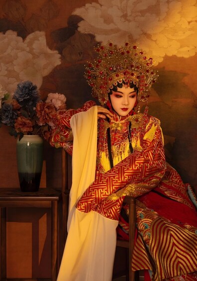 A person in traditional attire with a patterned robe and a decorative headdress, seated in a room with a floral arrangement and a vase on a table. 4