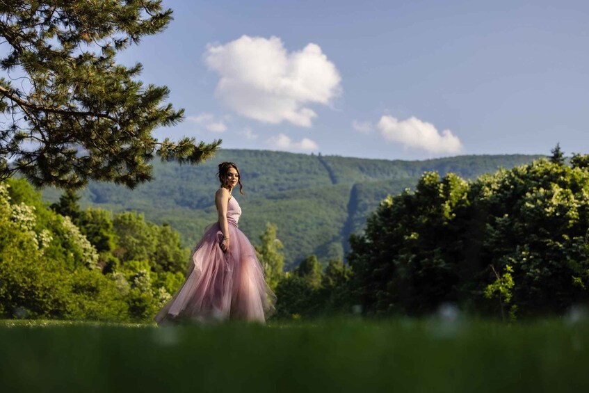 A person in a pink dress walking through a lush green field with trees and hills in the background. 5