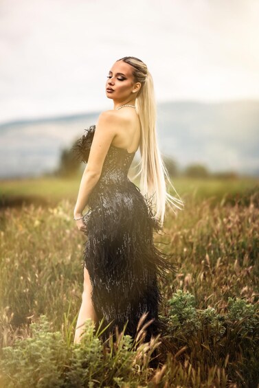A person in a black, feathered dress standing in a field with grass and distant hills. 3