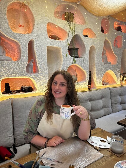 A person sitting at a table with a mug, surrounded by a wall with painted pottery shapes. 3