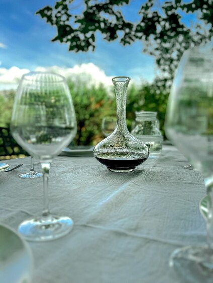 A table with a clear glass decanter, a wine glass, and a small glass jar, set outdoors with greenery in the background. 2