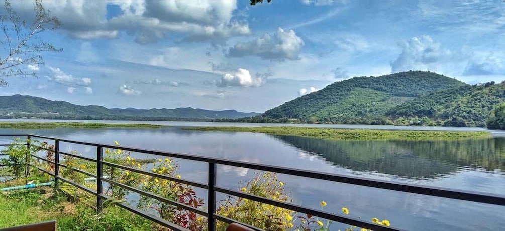 A calm lake with a metal railing in the foreground, surrounded by lush greenery and hills in the background. 2