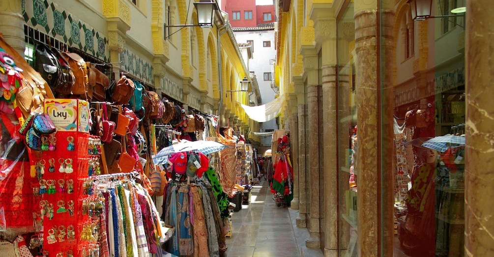 A narrow street with shops on both sides, displaying colorful bags and accessories. 2