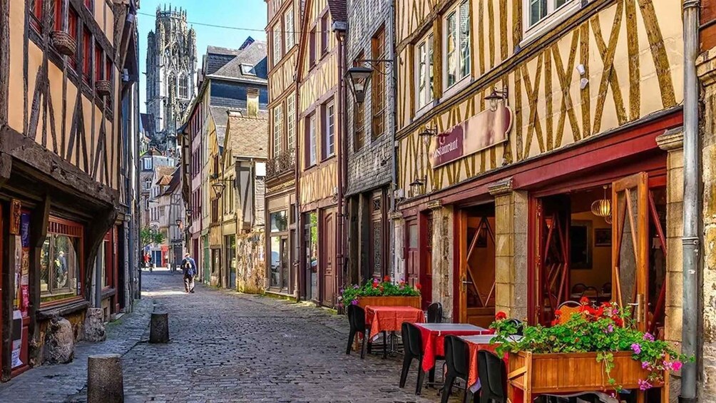 A cobblestone street lined with traditional half-timbered buildings, a church spire in the distance, and outdoor seating at a café. 3