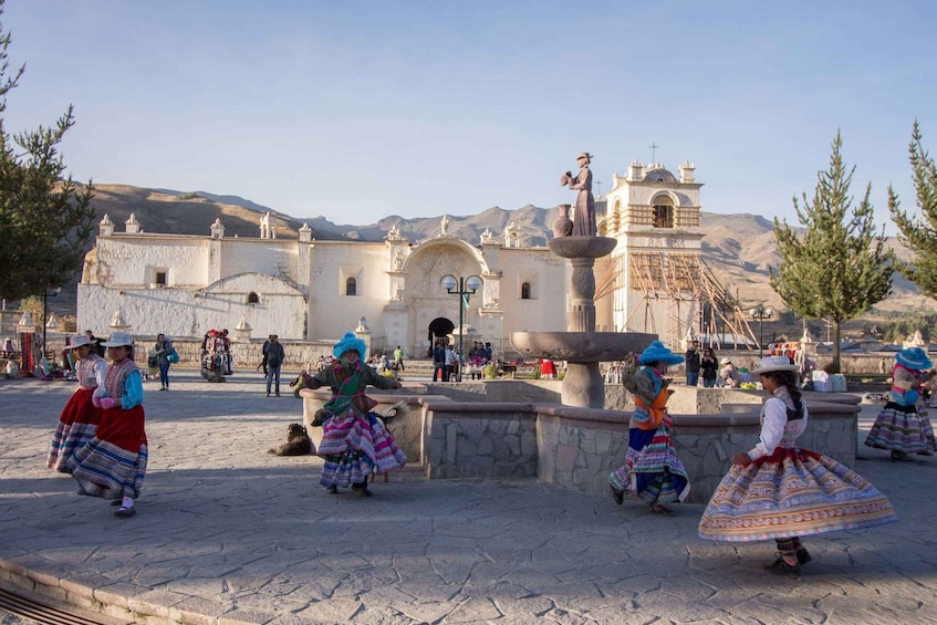 A plaza with people in traditional attire, a fountain, and a historic building with a bell tower. 4