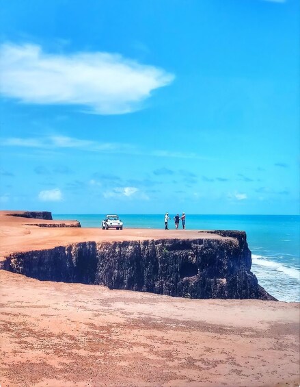 A beach with a white car parked on a cliff, three people standing near the edge, and the ocean in the background. 3