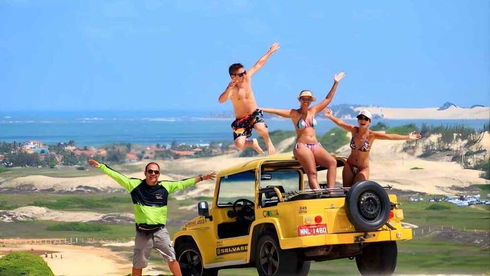 A yellow off-road vehicle with people standing on top, a person in a green shirt, and a beach landscape with dunes and the ocean in the background. 1