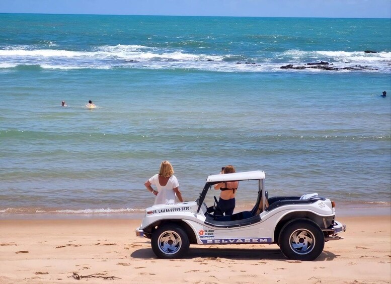 A beach with a white vehicle parked on the sand, a woman in a white top, and the ocean in the background. 1