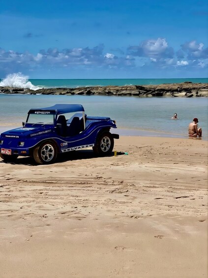 A blue off-road vehicle parked on a sandy beach with rocky cliffs and the ocean in the background. 4