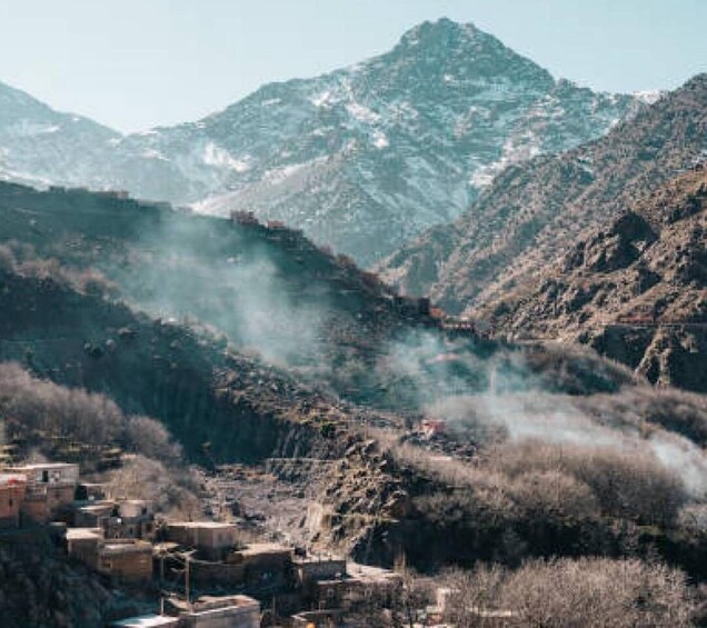 A mountainous landscape with a village at its base, smoke rising from the valley, and snow-capped peaks in the distance. 5