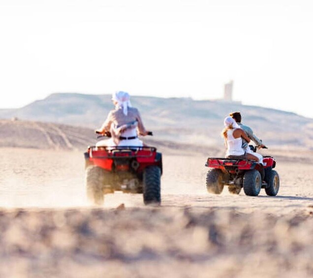 Two people riding ATVs on a desert terrain with a hill in the background. 2