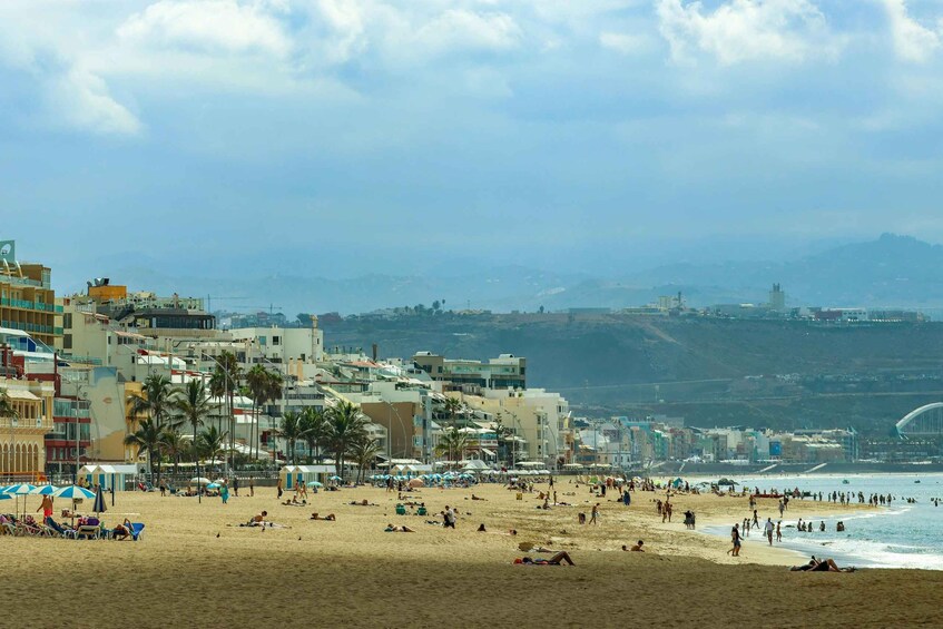 A crowded beach with people sunbathing and walking, palm trees, and a cityscape in the background. 5