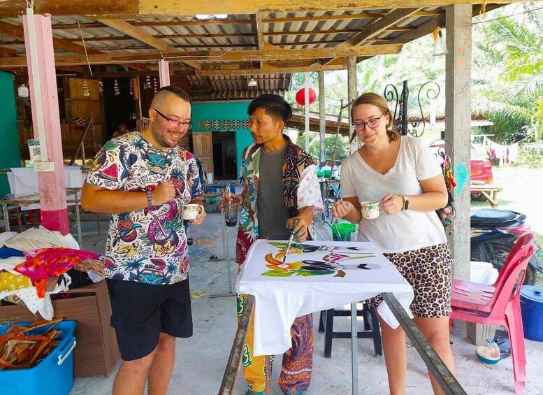Three people painting on a table under a covered area with a wooden ceiling and a green wall in the background. 5